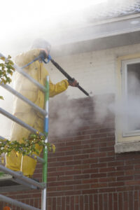 man in yellow rain suit cleans paint from brick wall of house facade with pressure washer while standing on scaffolding